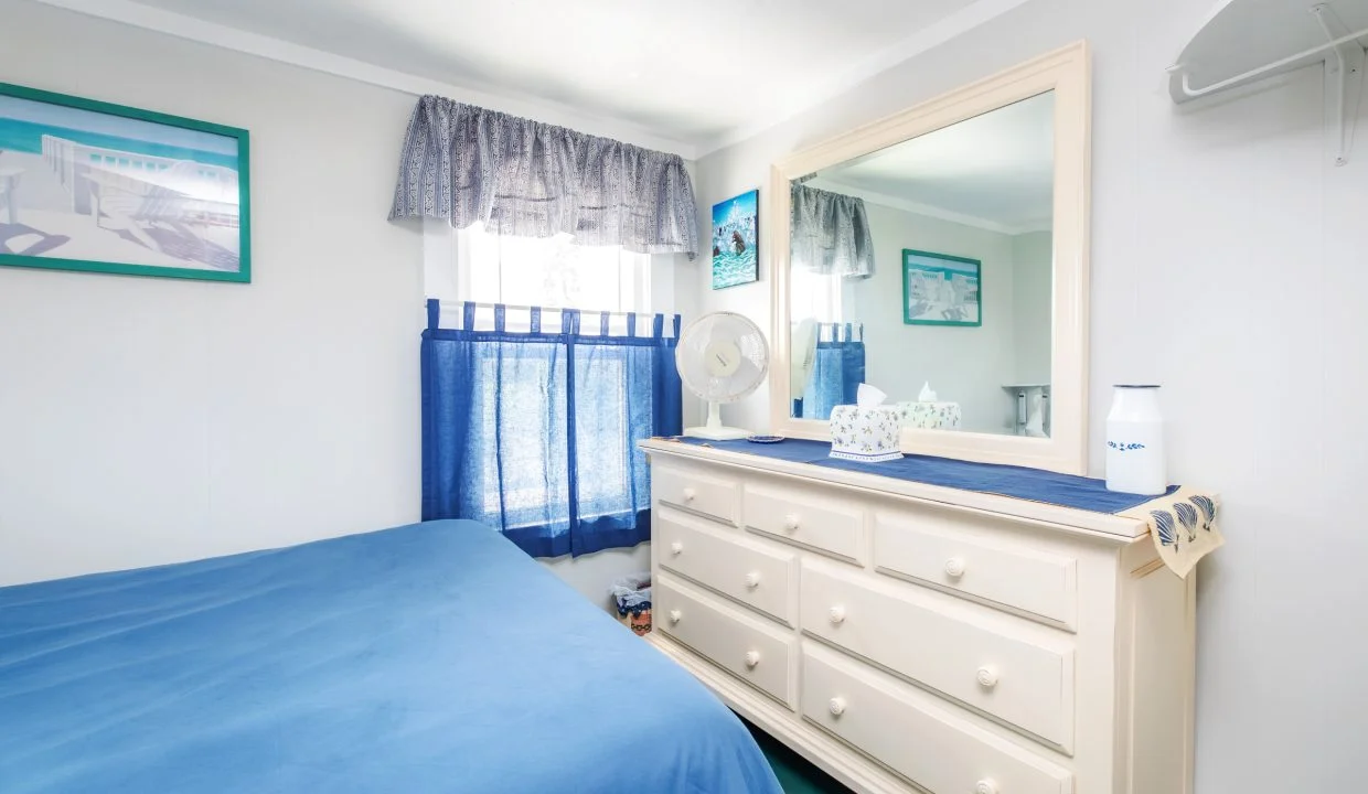 A small bedroom with white walls, a blue bedspread, a white dresser with a large mirror, and a window with blue curtains. The dresser has a white vase, a doily, and a fan. Artwork hangs on the walls.