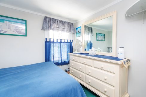 A small bedroom with white walls, a blue bedspread, a white dresser with a large mirror, and a window with blue curtains. The dresser has a white vase, a doily, and a fan. Artwork hangs on the walls.