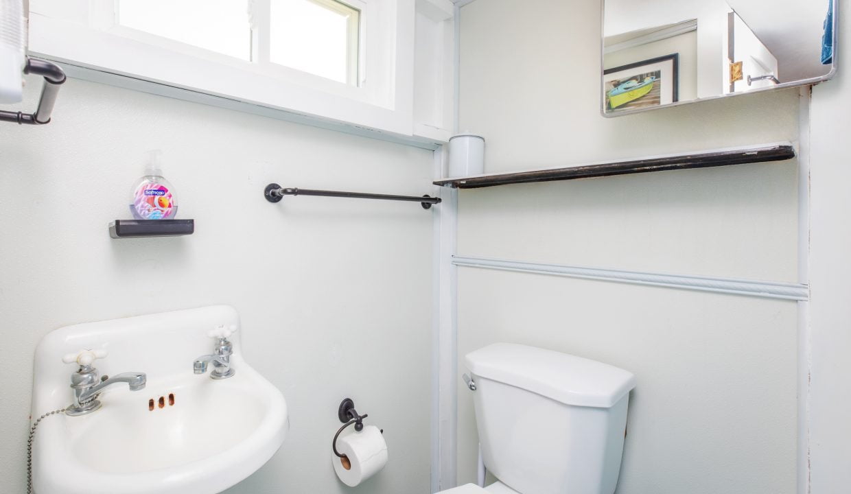 A small bathroom features a white sink, a toilet with a roll of toilet paper, and a wall-mounted shelf above the toilet. A window and mirror are also present.