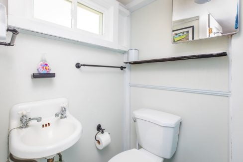 A small bathroom features a white sink, a toilet with a roll of toilet paper, and a wall-mounted shelf above the toilet. A window and mirror are also present.