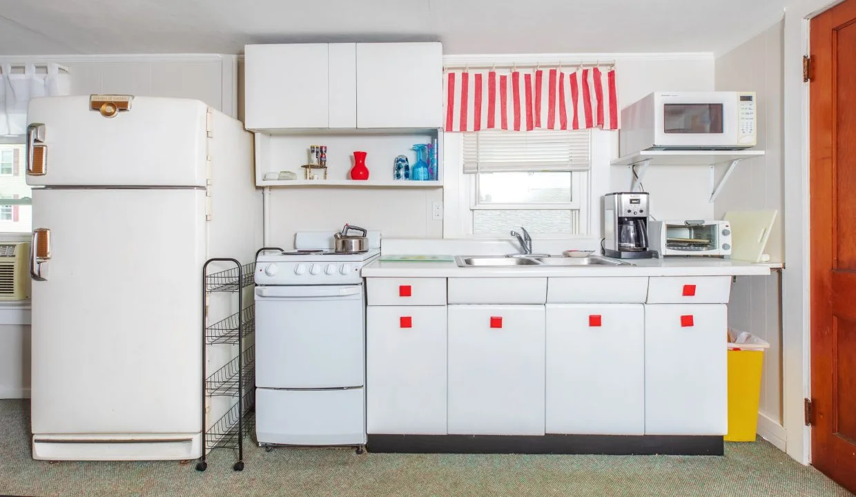 A compact kitchen features white cabinets, a small white stove, refrigerator, microwave, coffee maker, toaster, and striped red and white curtains over the sink window. Several appliances are on the countertop.