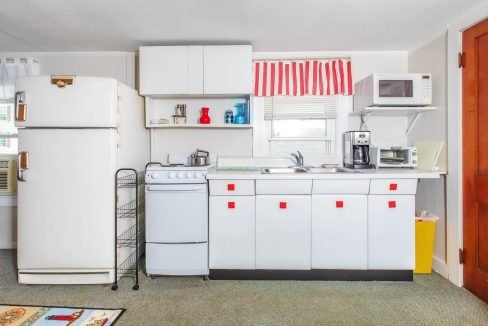 A compact kitchen features white cabinets, a small white stove, refrigerator, microwave, coffee maker, toaster, and striped red and white curtains over the sink window. Several appliances are on the countertop.