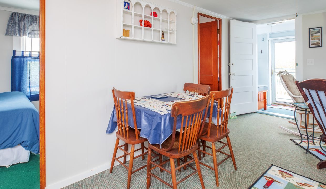 A dining area with a wooden table, four chairs, blue tablecloth, and wall-mounted shelf. A bedroom with a blue bed is seen to the left, and an open door leads to an enclosed porch on the right.