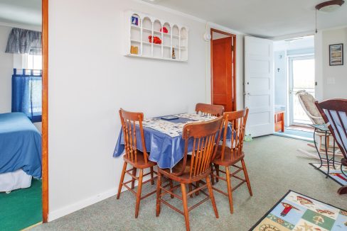 A dining area with a wooden table, four chairs, blue tablecloth, and wall-mounted shelf. A bedroom with a blue bed is seen to the left, and an open door leads to an enclosed porch on the right.