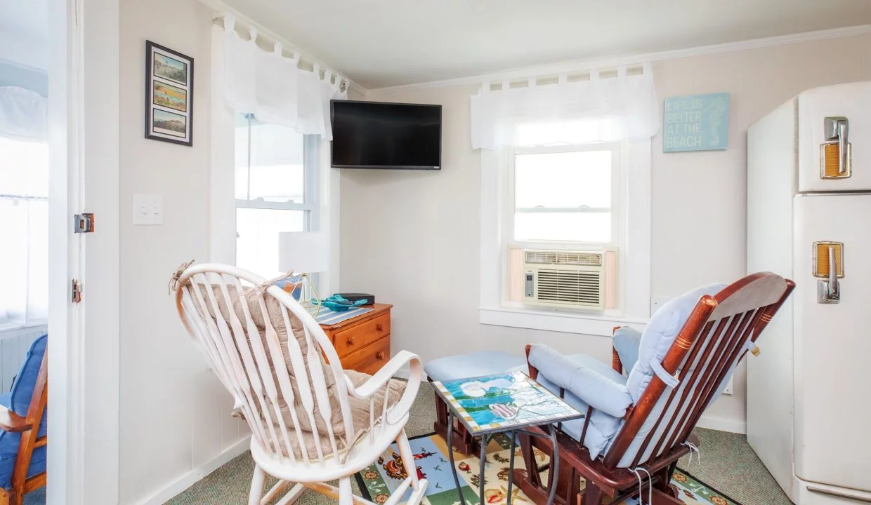 A small living room with a rocking chair, a cushioned chair, a table, a wall-mounted TV, an air conditioner in the window, and a vintage refrigerator against the wall.