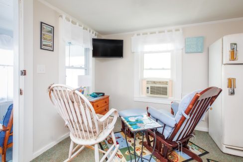 A small living room with a rocking chair, a cushioned chair, a table, a wall-mounted TV, an air conditioner in the window, and a vintage refrigerator against the wall.