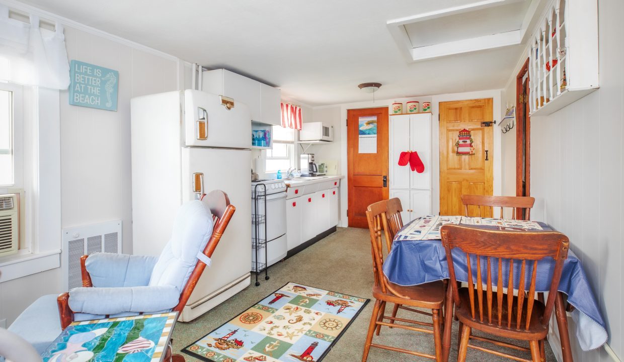 A small kitchen and dining area with a table and four chairs, a fridge, a stove, cabinets, and decorative items, including a seashell-themed rug and a sign on the wall.