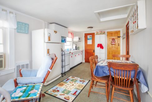 A small kitchen and dining area with a table and four chairs, a fridge, a stove, cabinets, and decorative items, including a seashell-themed rug and a sign on the wall.