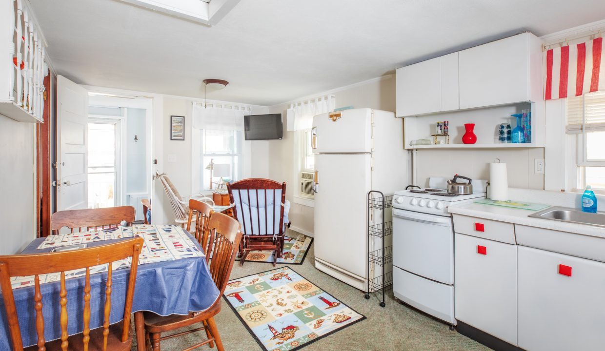 A compact kitchen and dining area with white cabinets, a refrigerator, a stove, a small table with chairs, and a rocking chair. Decor includes nautical-themed items and a small wall-mounted TV.