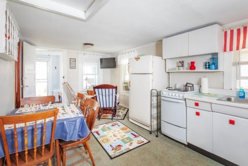 A compact kitchen and dining area with white cabinets, a refrigerator, a stove, a small table with chairs, and a rocking chair. Decor includes nautical-themed items and a small wall-mounted TV.