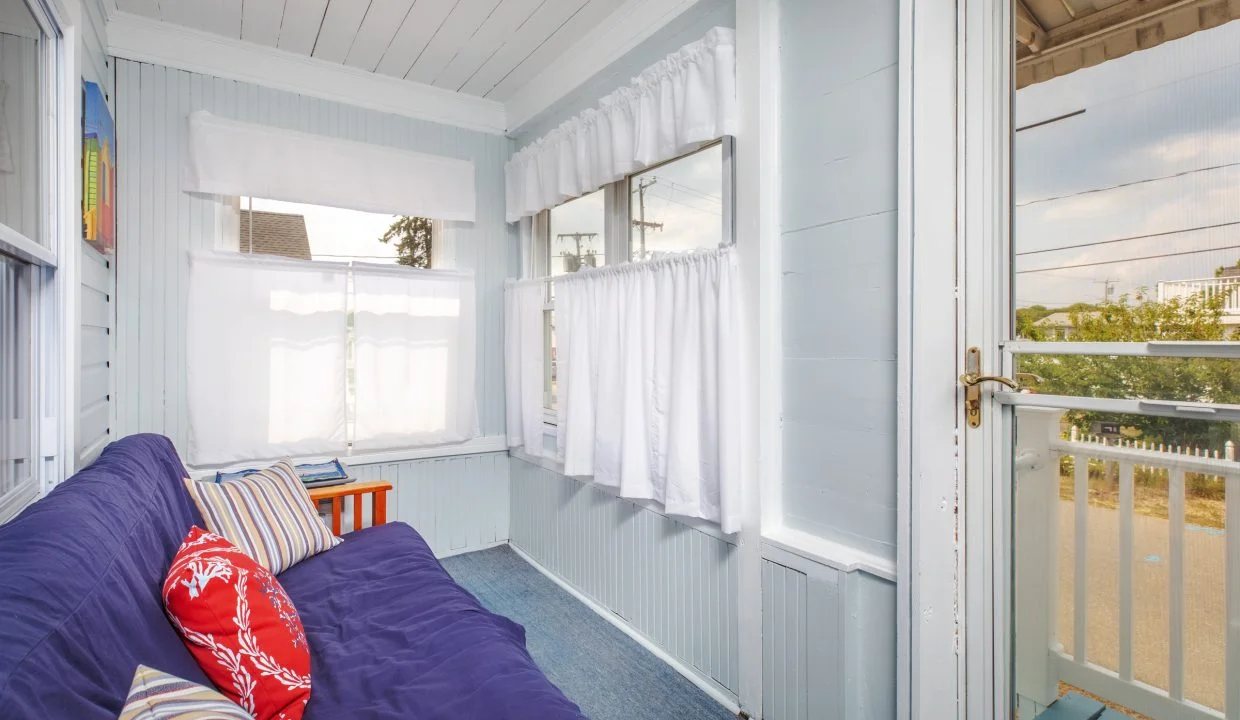 A cozy sunroom with light gray walls, multiple windows with white curtains, a blue futon with striped and patterned throw pillows, and a wooden side table.