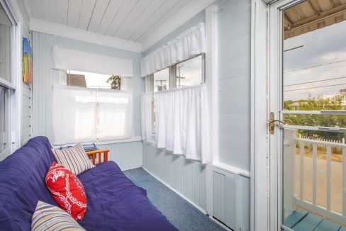 A cozy sunroom with light gray walls, multiple windows with white curtains, a blue futon with striped and patterned throw pillows, and a wooden side table.