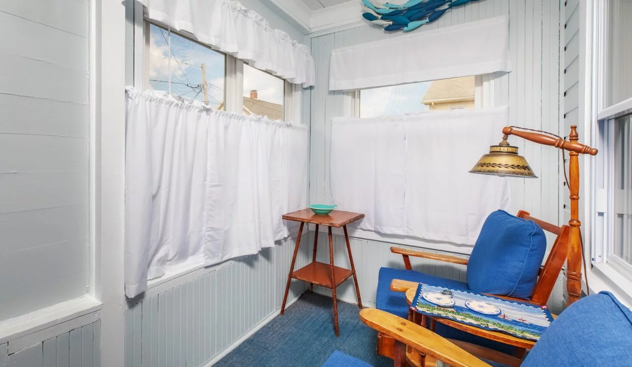 A small sunroom with light blue walls features two blue-cushioned wooden chairs, a wooden side table, and white curtains on the windows. The ceiling has white beadboard paneling and a fish wall decoration.