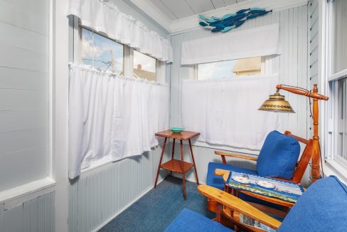 A small sunroom with light blue walls features two blue-cushioned wooden chairs, a wooden side table, and white curtains on the windows. The ceiling has white beadboard paneling and a fish wall decoration.