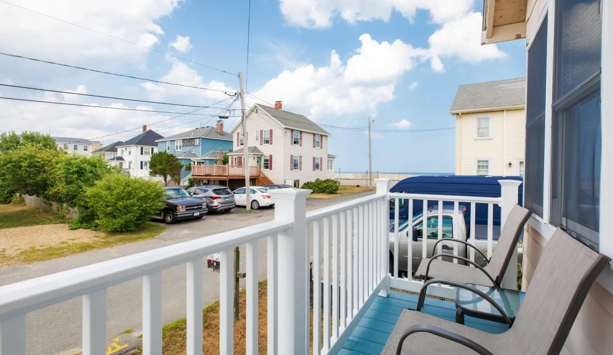 View from a porch with two chairs, overlooking a street with parked cars, houses, and a glimpse of the ocean in the background under a partly cloudy sky.