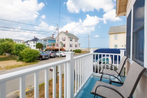 View from a porch with two chairs, overlooking a street with parked cars, houses, and a glimpse of the ocean in the background under a partly cloudy sky.