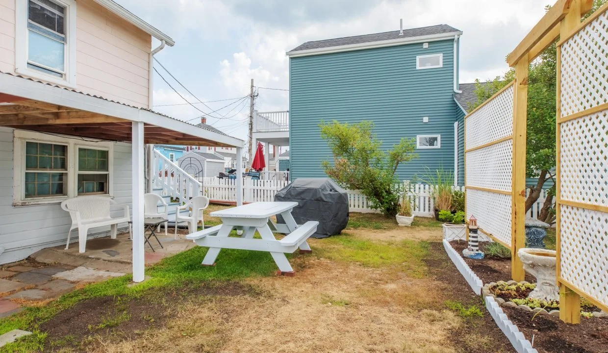 A small backyard with a picnic table, white chairs, a grill, a lattice screen with planters on the right, and adjacent houses in the background.