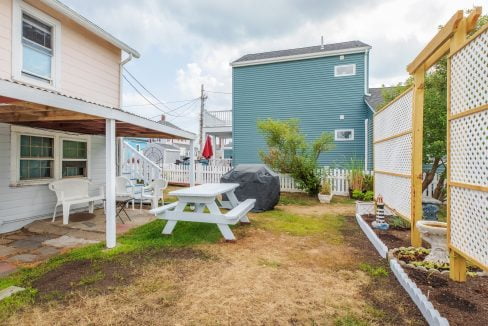 A small backyard with a picnic table, white chairs, a grill, a lattice screen with planters on the right, and adjacent houses in the background.