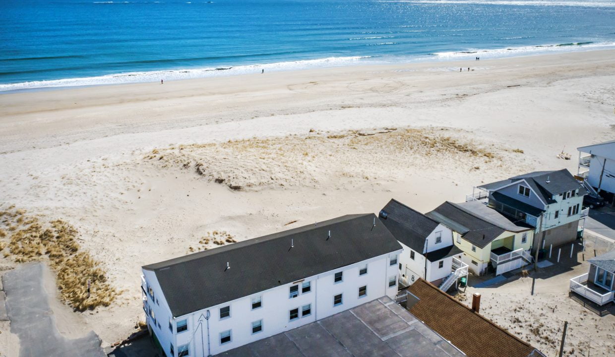 Aerial view of a coastline with sandy beach and few houses near the shoreline, under a clear blue sky with calm ocean waves.