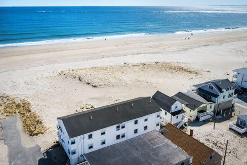 Aerial view of a coastline with sandy beach and few houses near the shoreline, under a clear blue sky with calm ocean waves.