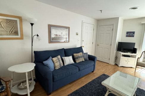 A living room with a blue couch, white side table, wicker coffee table, floor lamp, wall art, TV on a stand, and a dark blue rug on a wooden floor.