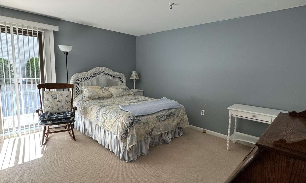 A bedroom with a bed, a nightstand, a wooden dresser, a rocking chair, and a standing lamp. Sliding glass doors with vertical blinds allow natural light into the room. Walls are painted gray.