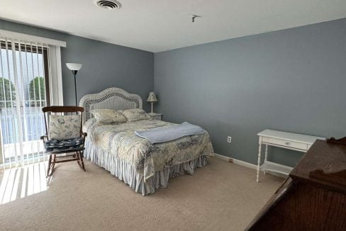 A bedroom with a bed, a nightstand, a wooden dresser, a rocking chair, and a standing lamp. Sliding glass doors with vertical blinds allow natural light into the room. Walls are painted gray.