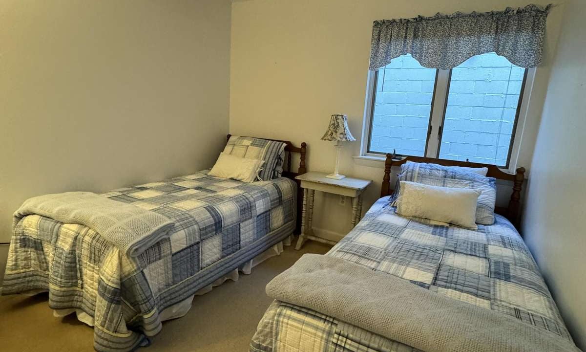 A small bedroom with two single beds, each covered with checkered quilts. A small table with a lamp is between the beds. The room has a window with a blue valance curtain above it.