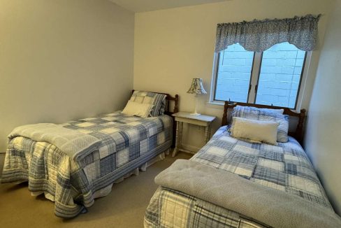 A small bedroom with two single beds, each covered with checkered quilts. A small table with a lamp is between the beds. The room has a window with a blue valance curtain above it.