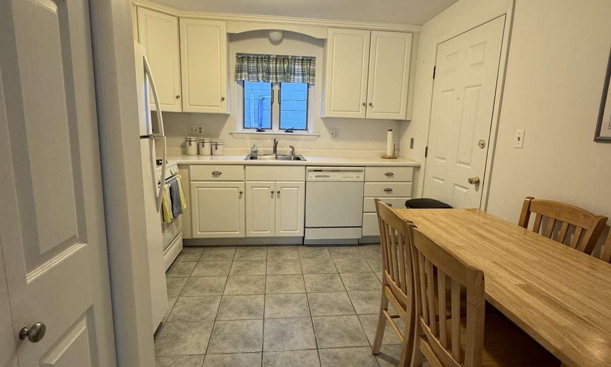 A small kitchen with white cabinets, a tiled floor, a wooden table with four chairs, a refrigerator, stove, dishwasher, and a window above the sink.