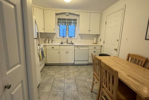 A small kitchen with white cabinets, a tiled floor, a wooden table with four chairs, a refrigerator, stove, dishwasher, and a window above the sink.