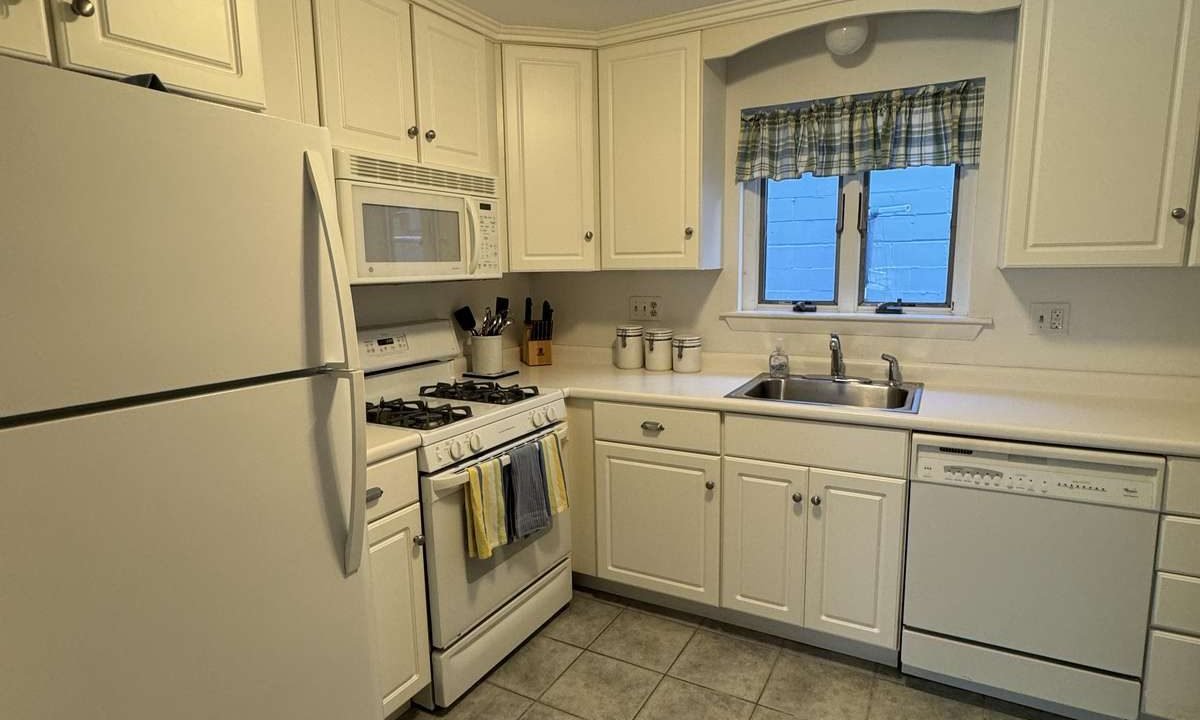 A small kitchen with white cabinets, a white refrigerator, gas stove, microwave, and dishwasher. The countertop is white, and the floor is covered with gray tiles. A window with a plaid curtain is above the sink.