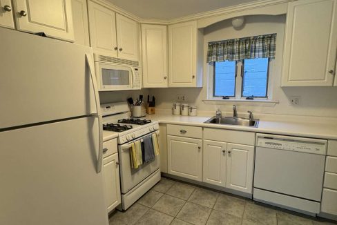 A small kitchen with white cabinets, a white refrigerator, gas stove, microwave, and dishwasher. The countertop is white, and the floor is covered with gray tiles. A window with a plaid curtain is above the sink.