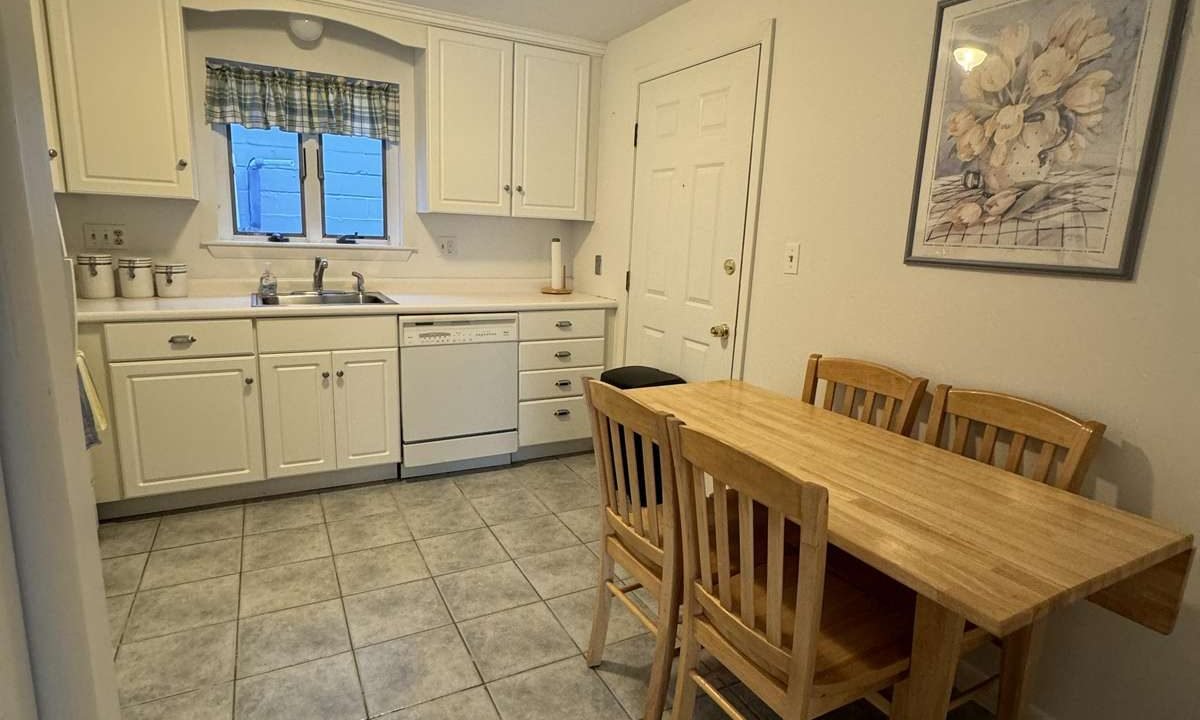 A small kitchen with white cabinets, a dishwasher, and a sink under a window. There's a wooden table with three chairs and a flower painting on the wall. The floor is tiled.