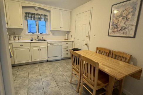 A small kitchen with white cabinets, a dishwasher, and a sink under a window. There's a wooden table with three chairs and a flower painting on the wall. The floor is tiled.