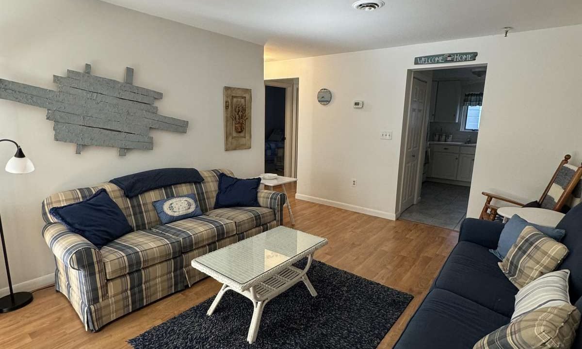 A living room with two sofas, a blue rug, a coffee table, a standing lamp, and wall decor. A hallway and bathroom entrance are visible in the background.