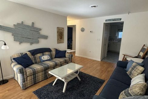 A living room with two sofas, a blue rug, a coffee table, a standing lamp, and wall decor. A hallway and bathroom entrance are visible in the background.