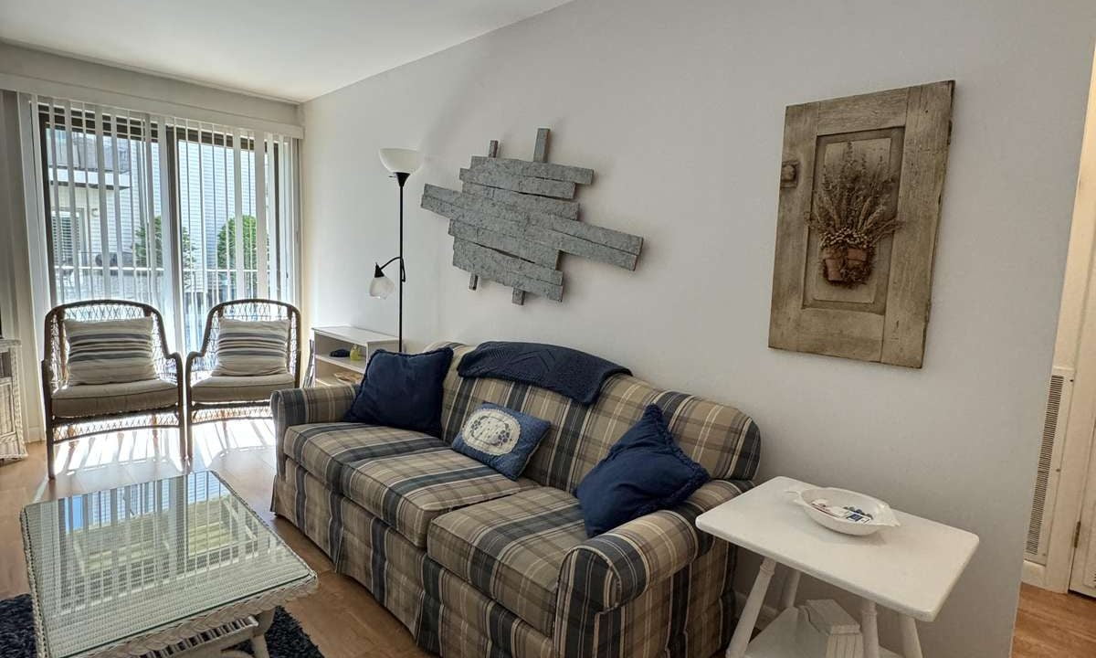 A cozy living room features a plaid sofa with blue and white cushions, a glass-top coffee table, two wicker chairs near a window with vertical blinds, and nautical decor on the walls.