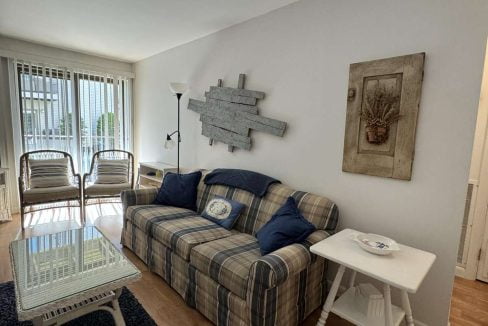 A cozy living room features a plaid sofa with blue and white cushions, a glass-top coffee table, two wicker chairs near a window with vertical blinds, and nautical decor on the walls.