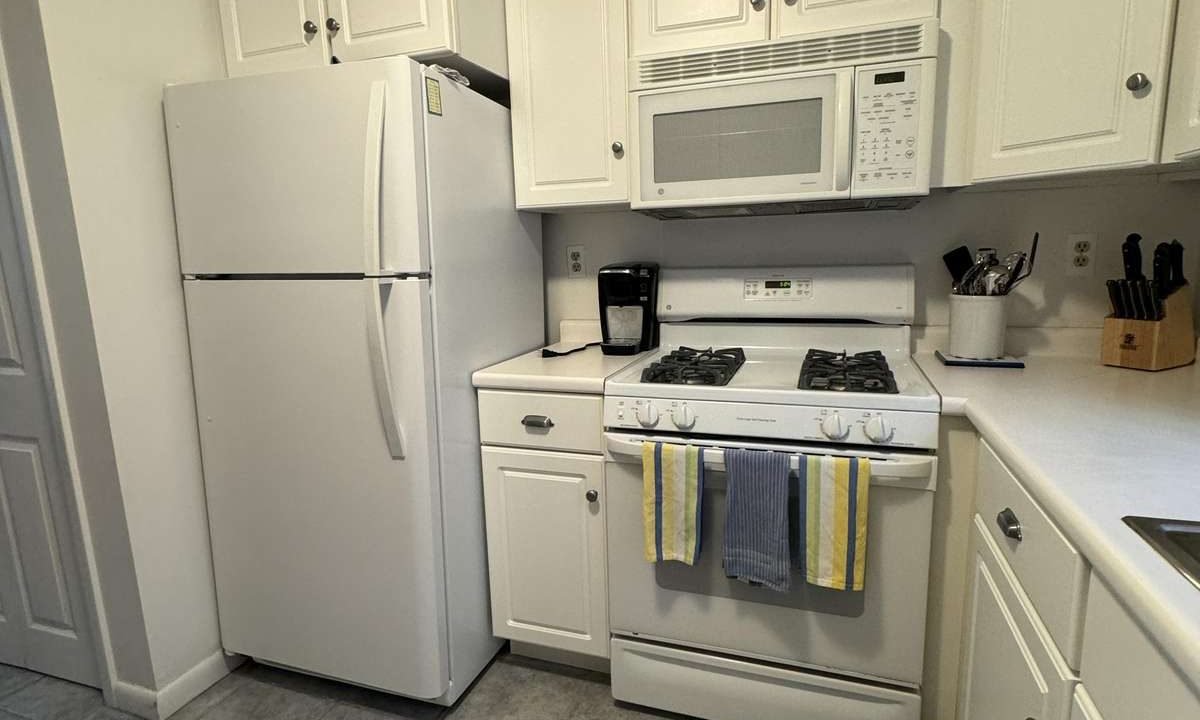 A kitchen with white appliances, including a refrigerator and a stove with an overhead microwave. White cabinets, a coffee maker, and utensils are visible. The floor is tiled in gray.