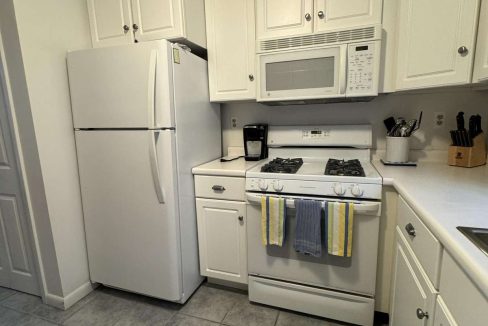 A kitchen with white appliances, including a refrigerator and a stove with an overhead microwave. White cabinets, a coffee maker, and utensils are visible. The floor is tiled in gray.