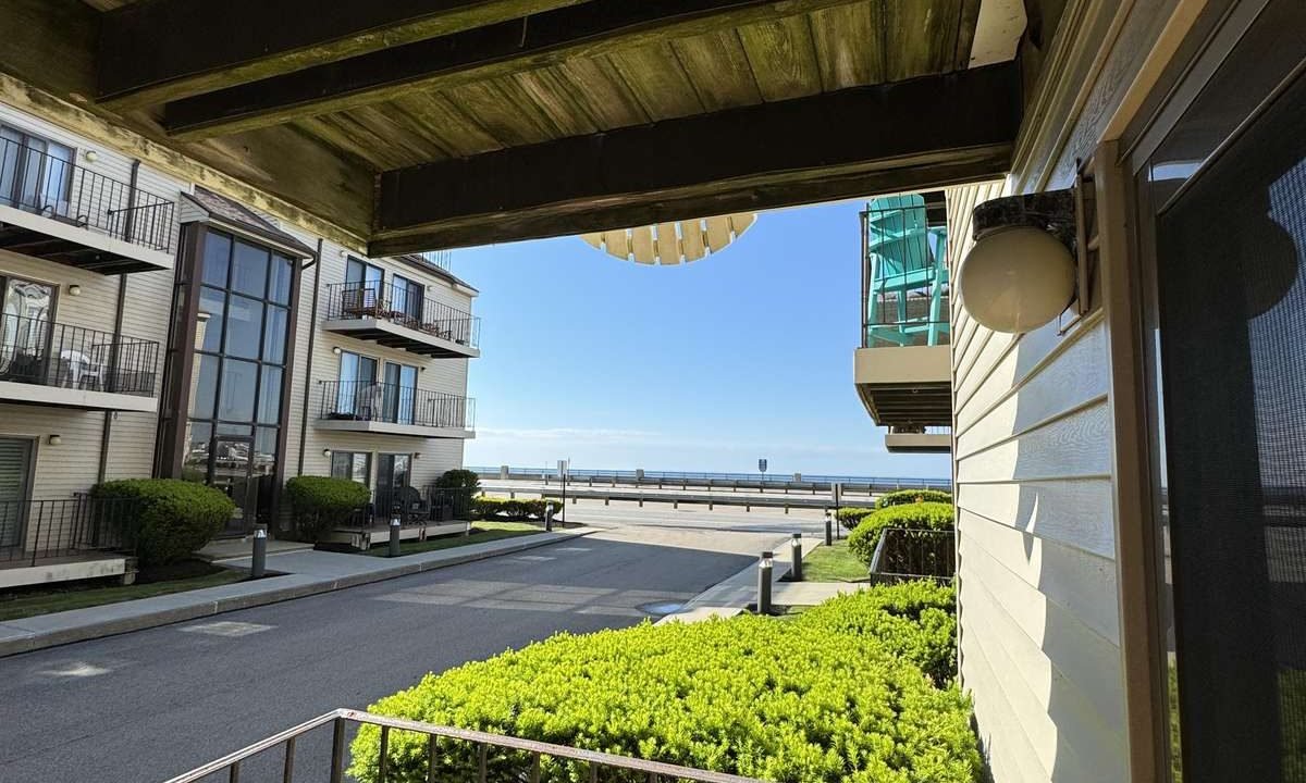 A view from a balcony overlooking a street lined with buildings, green bushes along the sidewalk, and a clear blue sky in the background.