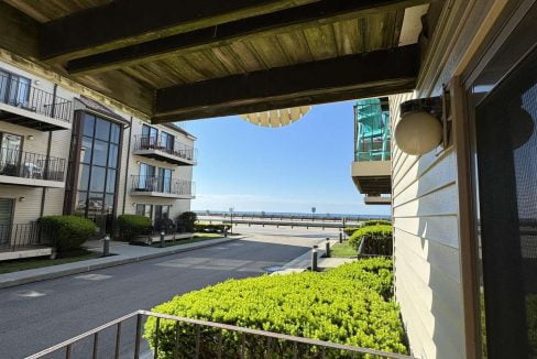 A view from a balcony overlooking a street lined with buildings, green bushes along the sidewalk, and a clear blue sky in the background.