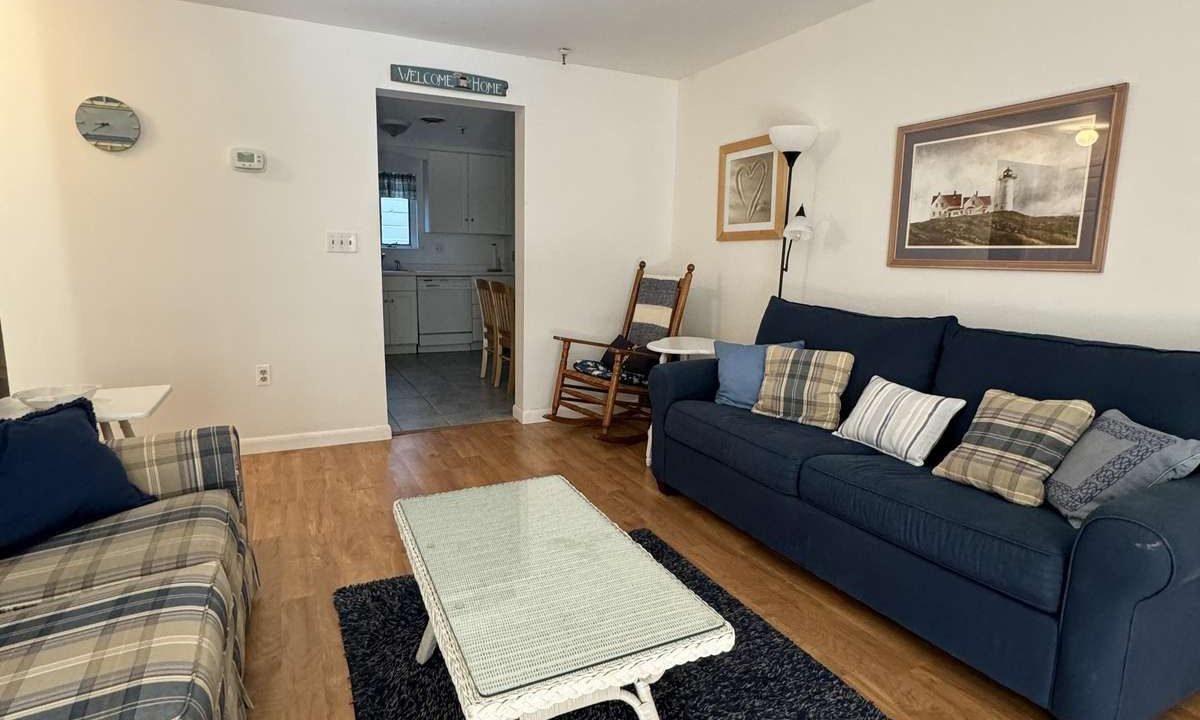 A living room with blue and plaid sofas, a white wicker coffee table, a blue rug, wooden floor, and a view into a small kitchen. The room has white walls with framed pictures and a standing lamp.