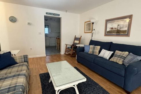 A living room with blue and plaid sofas, a white wicker coffee table, a blue rug, wooden floor, and a view into a small kitchen. The room has white walls with framed pictures and a standing lamp.