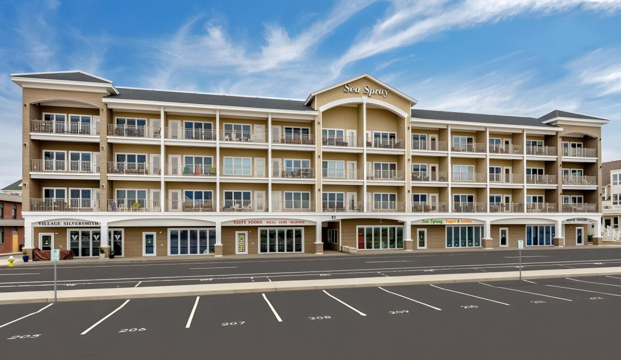 Four-story beige building named Sea Spray with shops on the ground floor and balconies on upper levels. Empty parking lot in foreground; blue sky with few clouds in background.