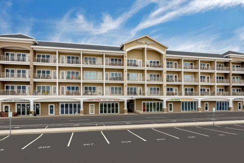 Four-story beige building named Sea Spray with shops on the ground floor and balconies on upper levels. Empty parking lot in foreground; blue sky with few clouds in background.