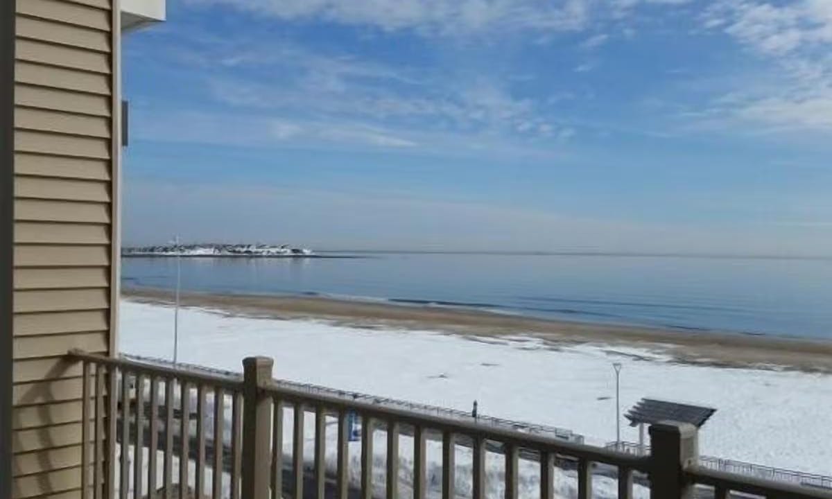 Balcony view of a beach with snowy sand and calm water under a partly cloudy blue sky. A building corner and railing are visible in the foreground.