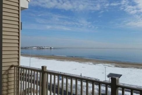 Balcony view of a beach with snowy sand and calm water under a partly cloudy blue sky. A building corner and railing are visible in the foreground.