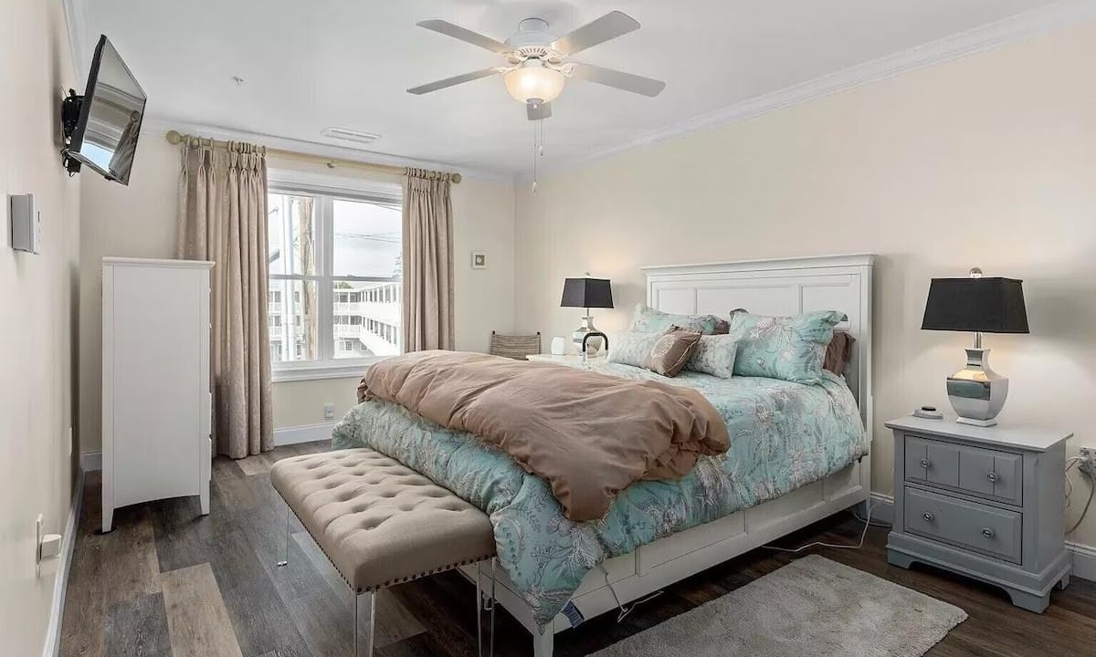 A bedroom with a double bed, floral bedding, a bench at the foot, a ceiling fan, a wall-mounted TV, and a window with curtains. Two nightstands with lamps are beside the bed.